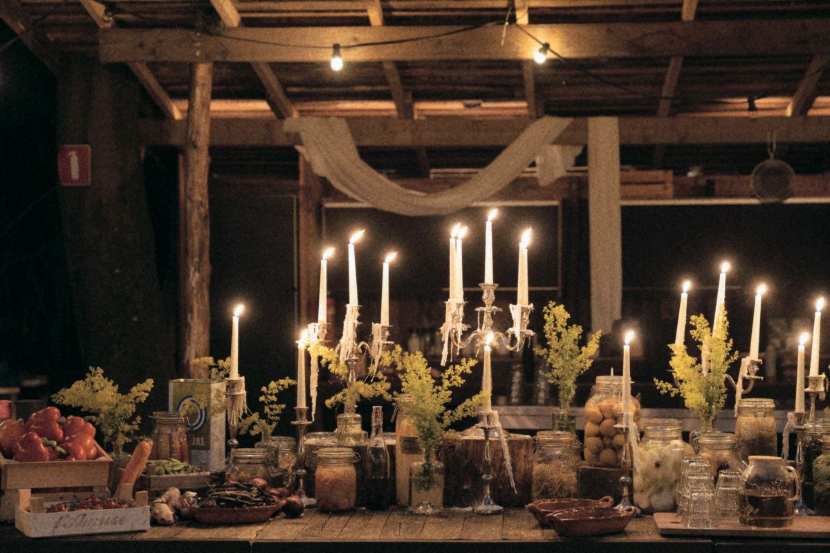Candles and jars of preserves on a long table in a wooden barn at night.
